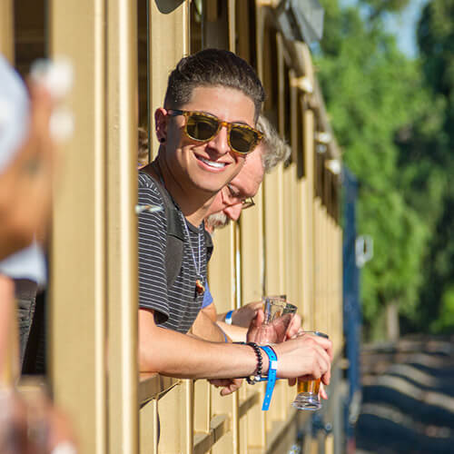 A smiling man leans out of a train car holding a beer