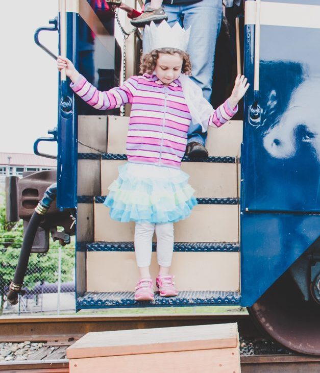 A young girl in a princess costume disembarks a blue train car