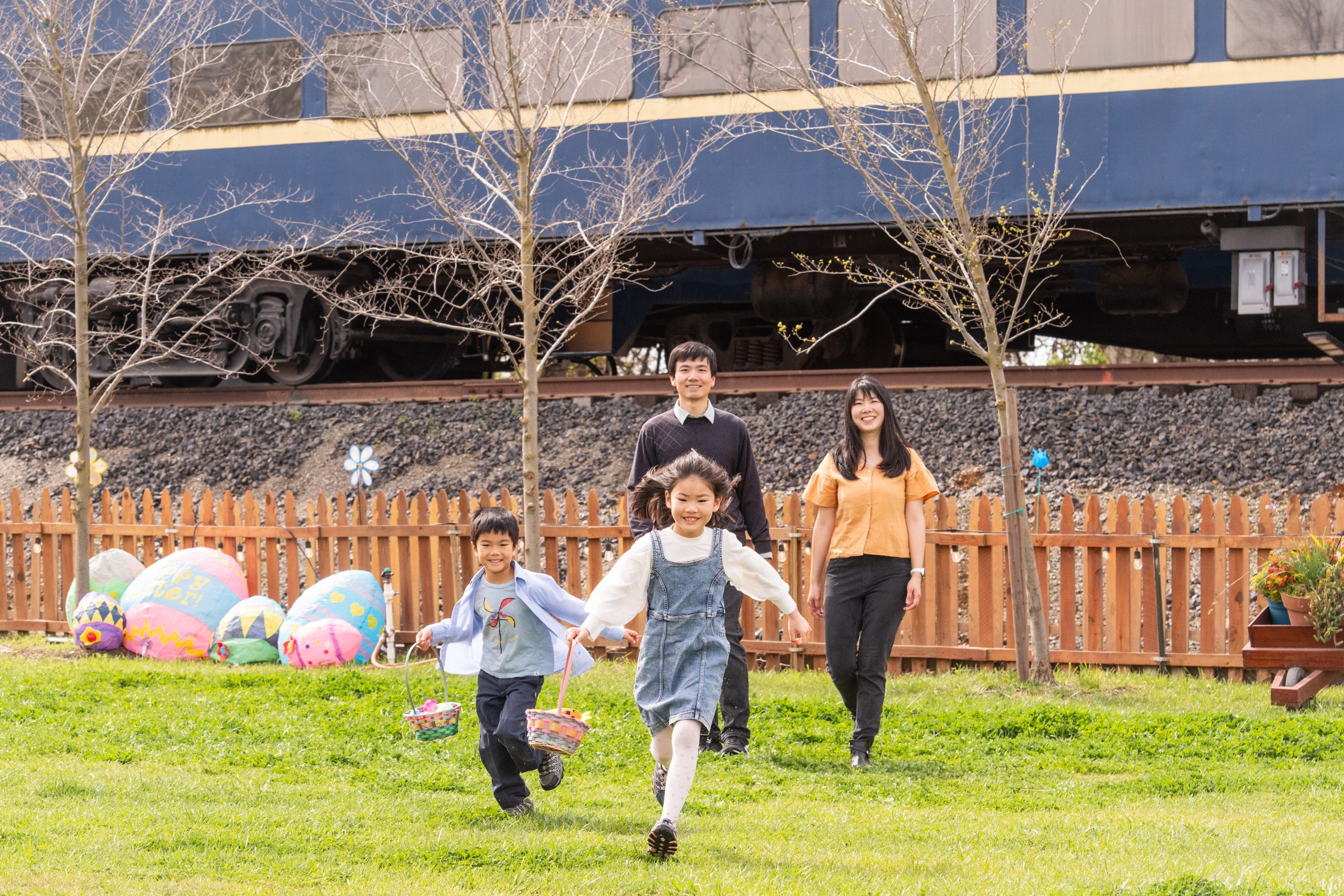 Family with two kids running on grass near painted eggs and train.