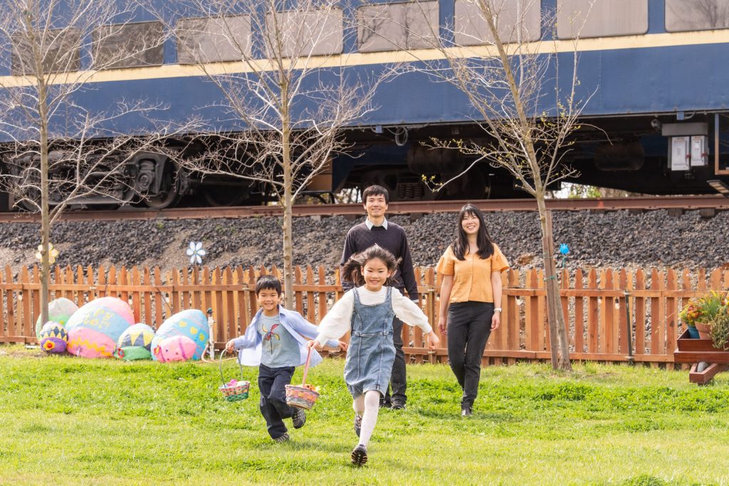Family with two kids running on grass near painted eggs and train.