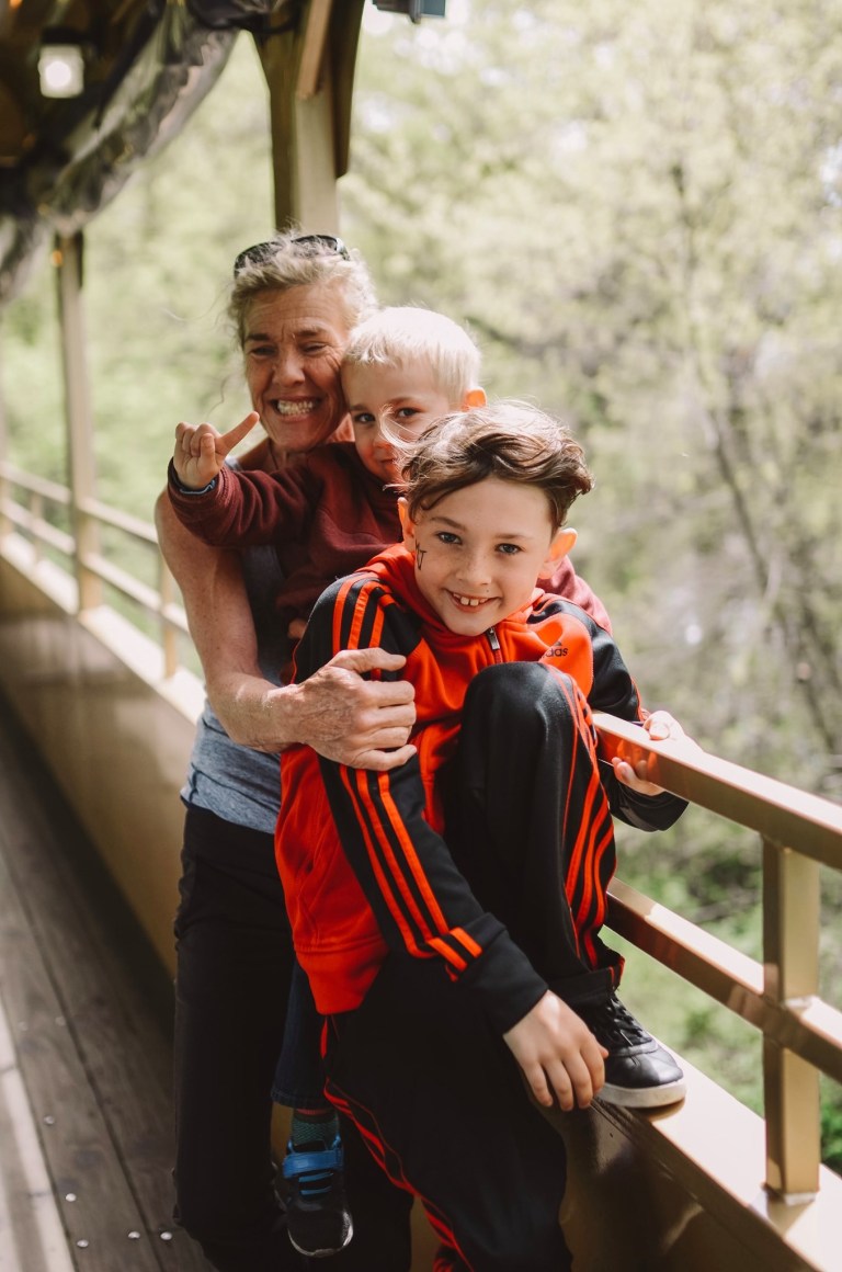 Smiling woman hugs two children on a wooden bridge.