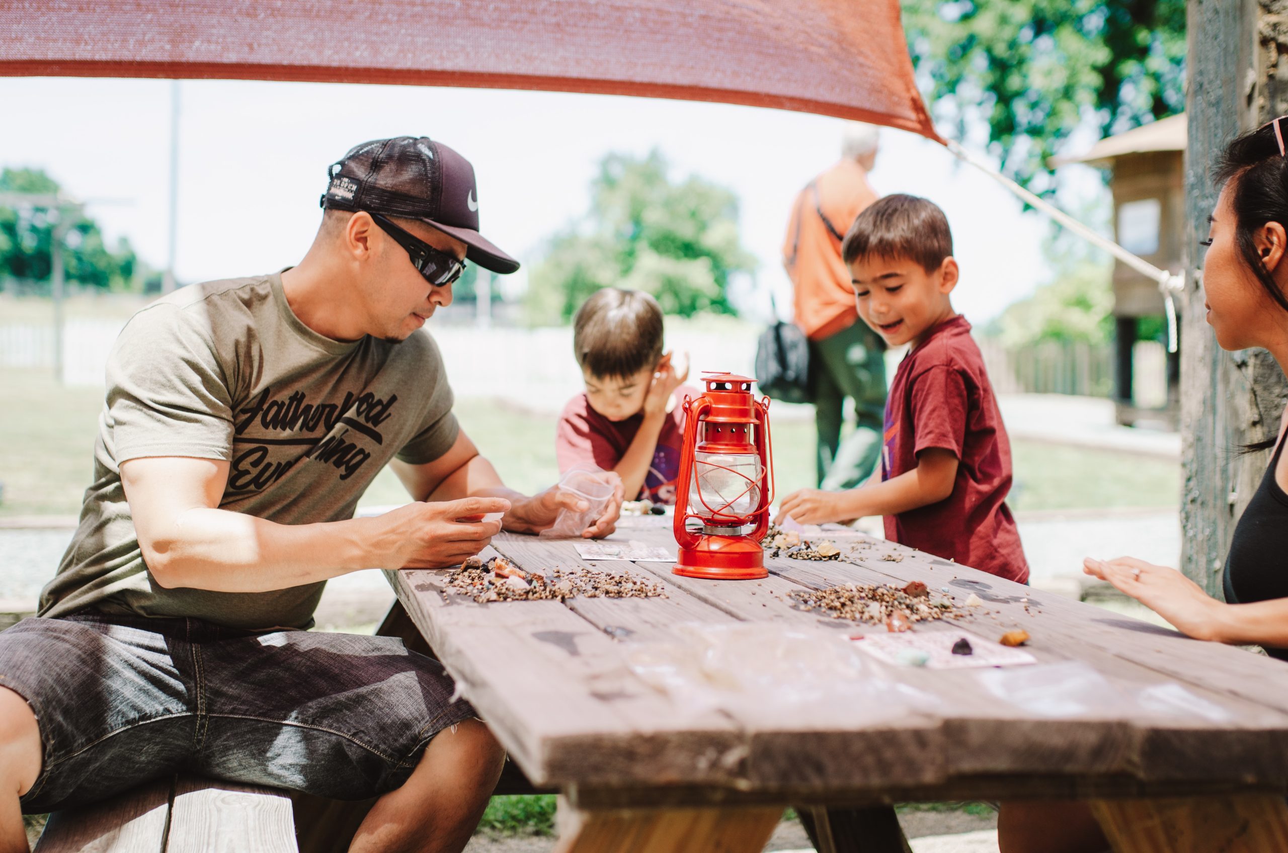 Family crafting with beads at a picnic table outdoors under a canopy