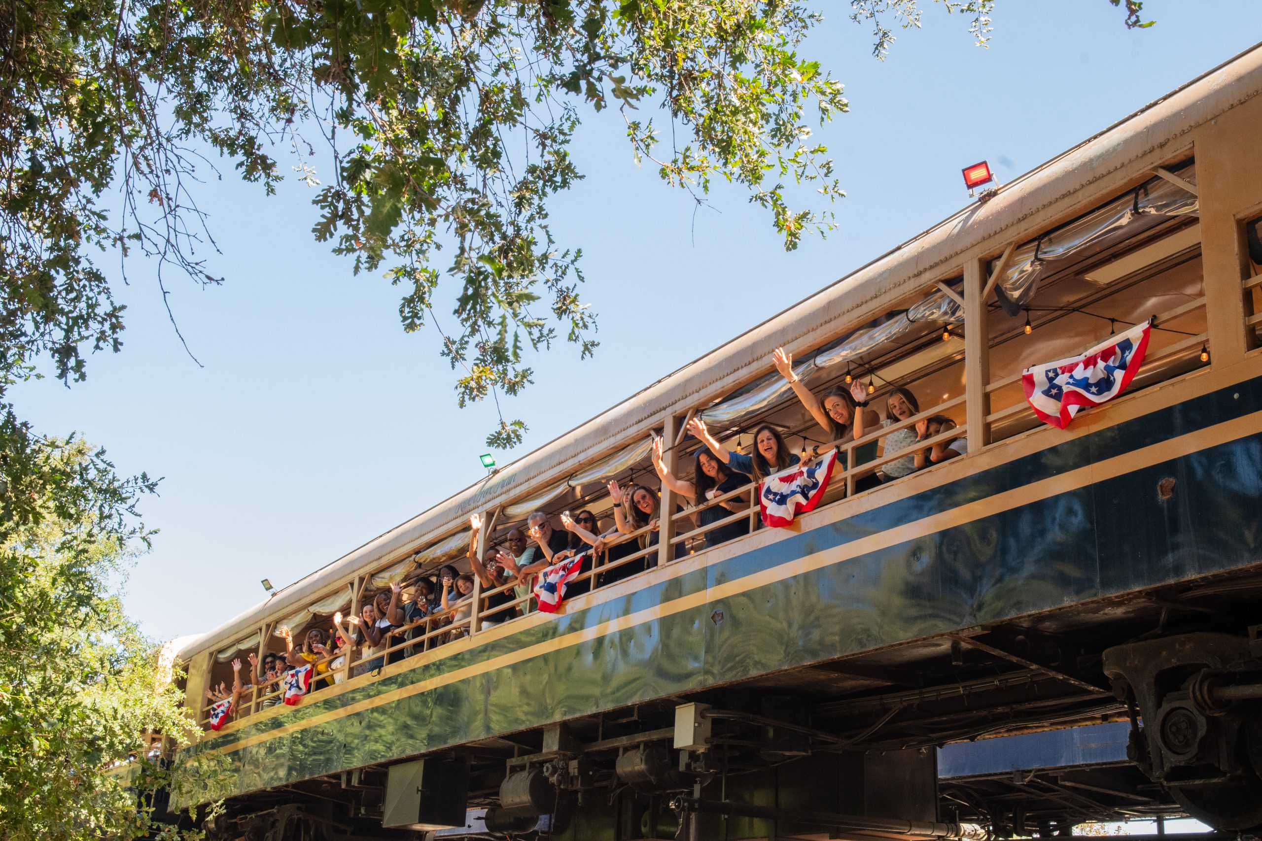People wave from train decorated with red, white, and blue banners under a clear sky.