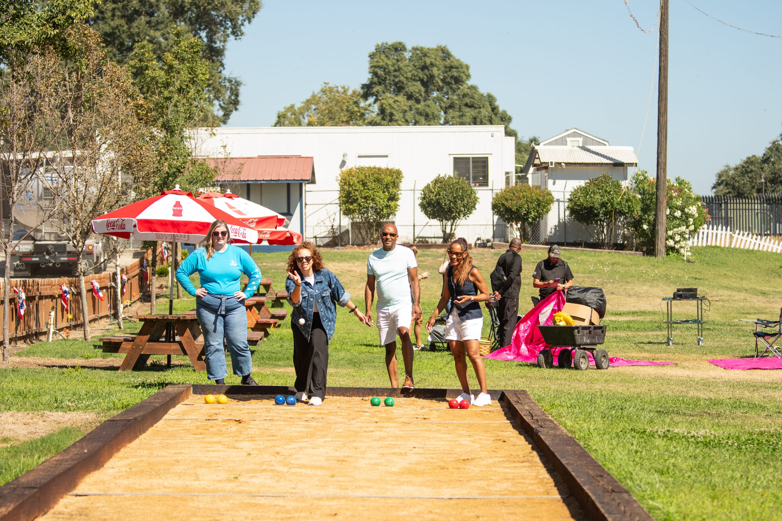Group playing bocce outdoors on a sunny day with picnic tables nearby.