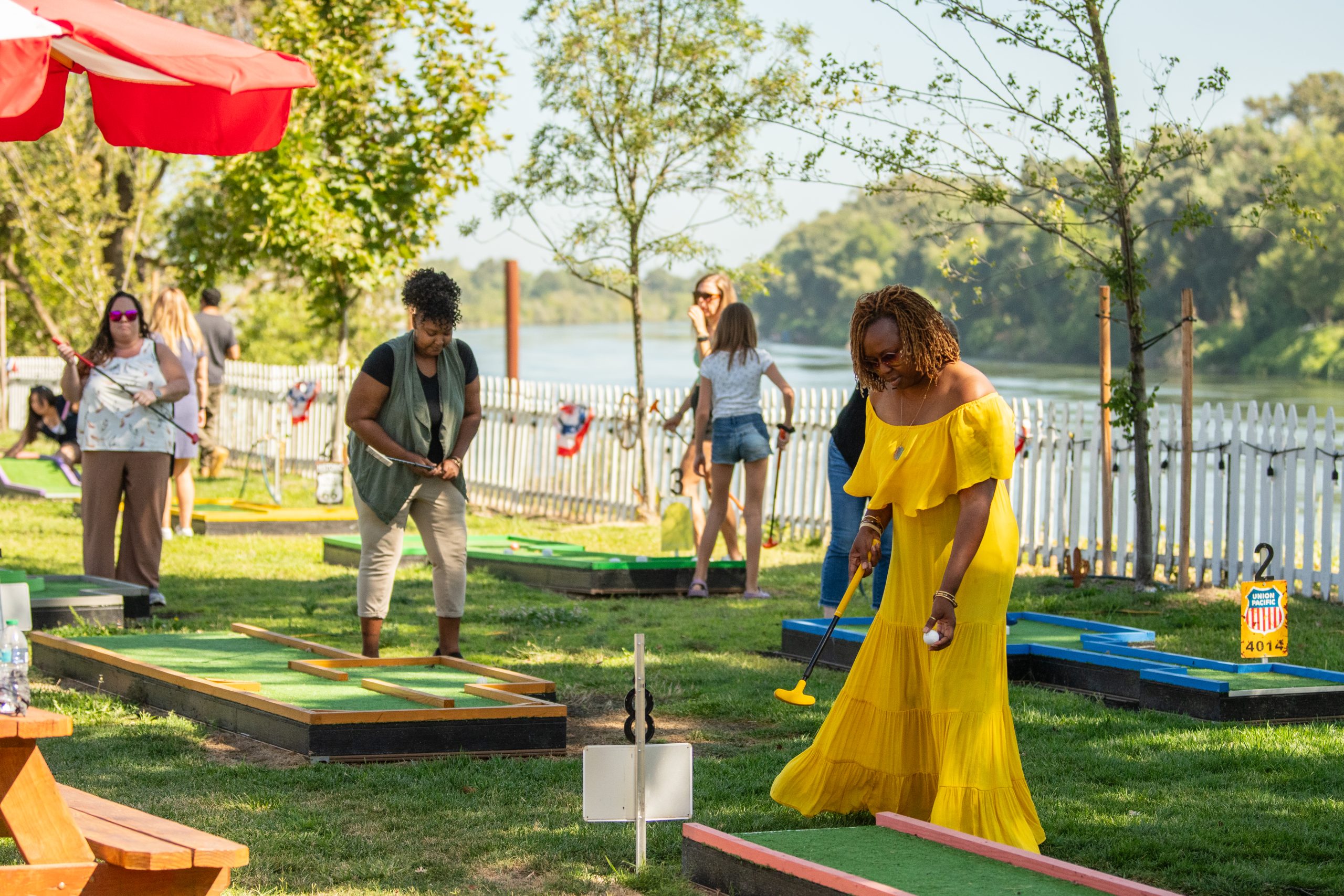 People play mini-golf outdoors near a river on a sunny day.