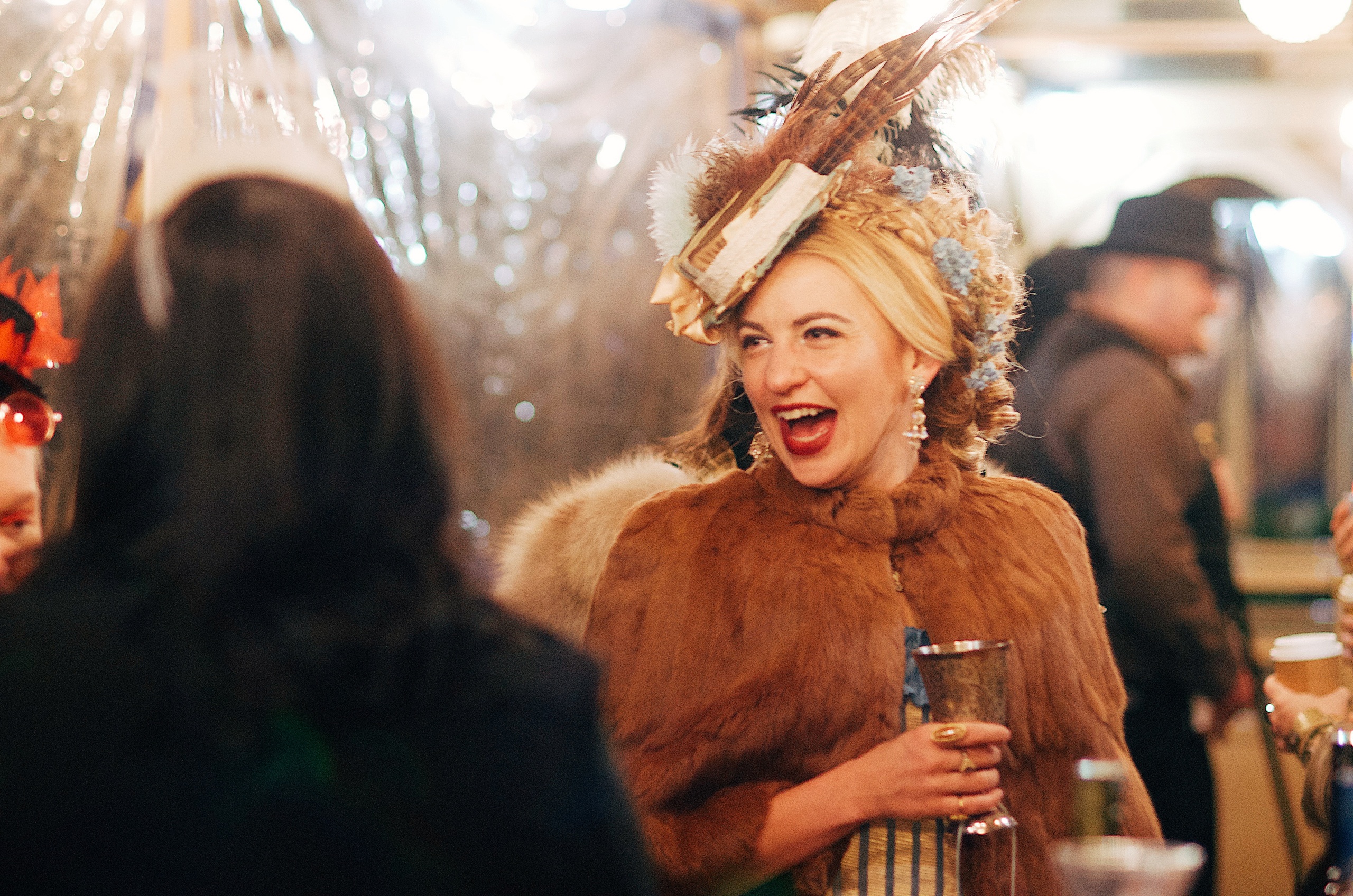 Woman in fur coat and feathered hat laughs at an event.