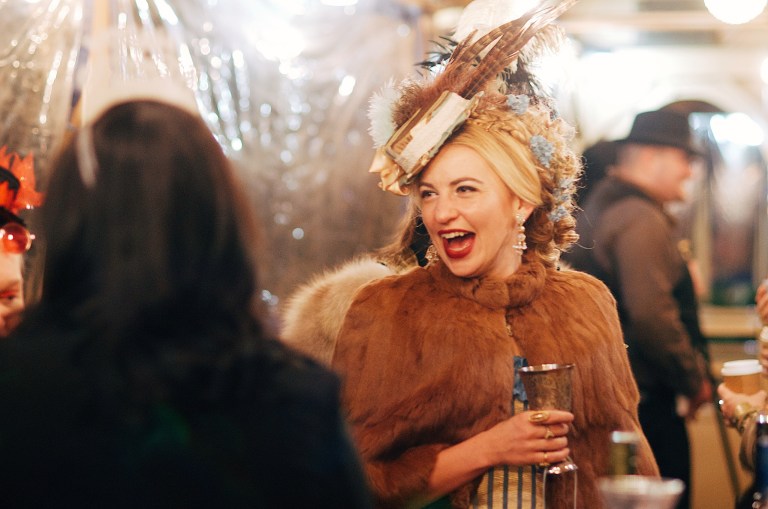 Woman in fur coat and feathered hat laughs at an event.