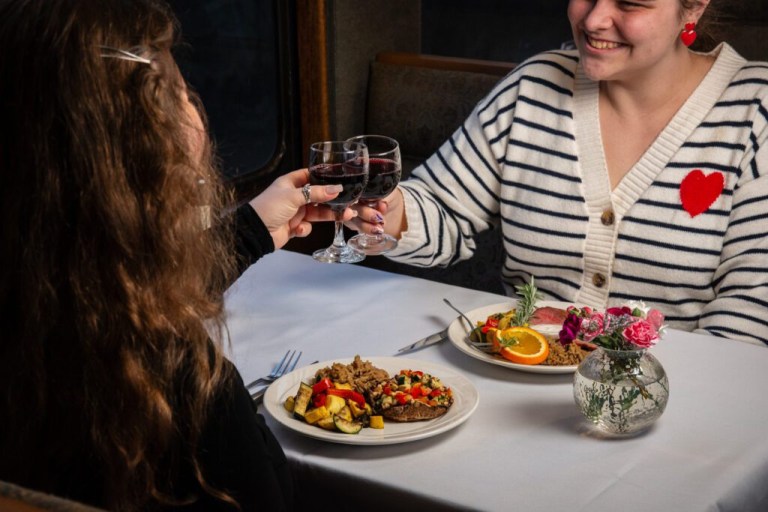 Two people clinking glasses over meals at a table, smiling.