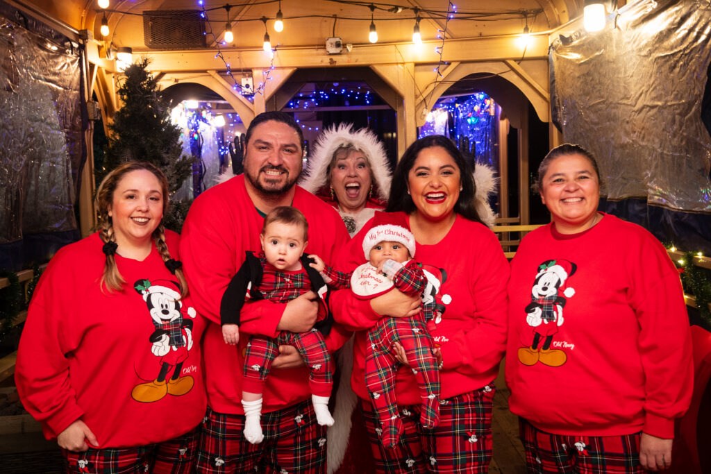 Family in matching holiday pajamas, two holding babies, joyful expressions, festive lights in background.