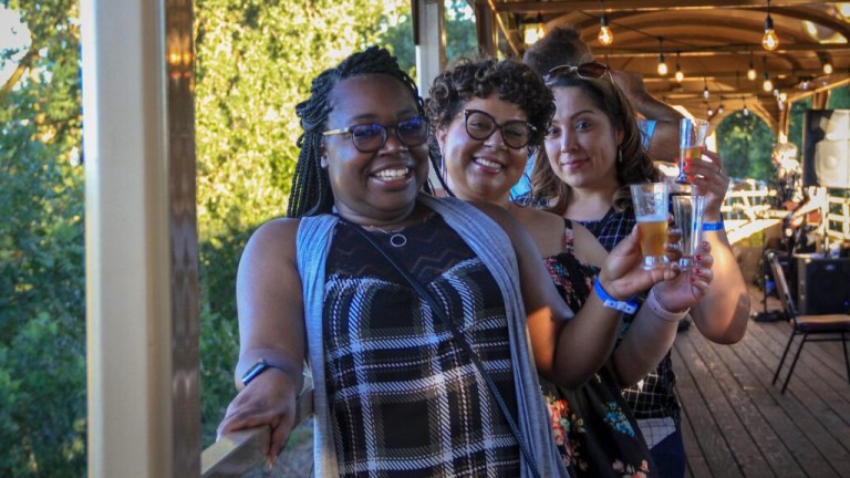 Three smiling women toasting with drinks on a sunny deck.