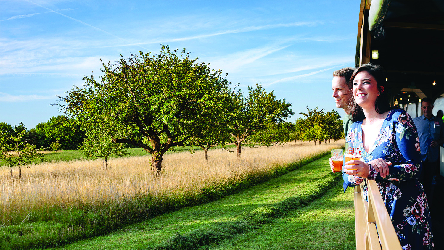 Two people with drinks enjoying a scenic view from a train with grassy fields and trees.