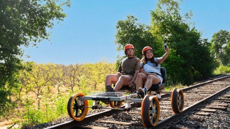 Two people on rail bike with orange wheels, taking selfie on sunny day.