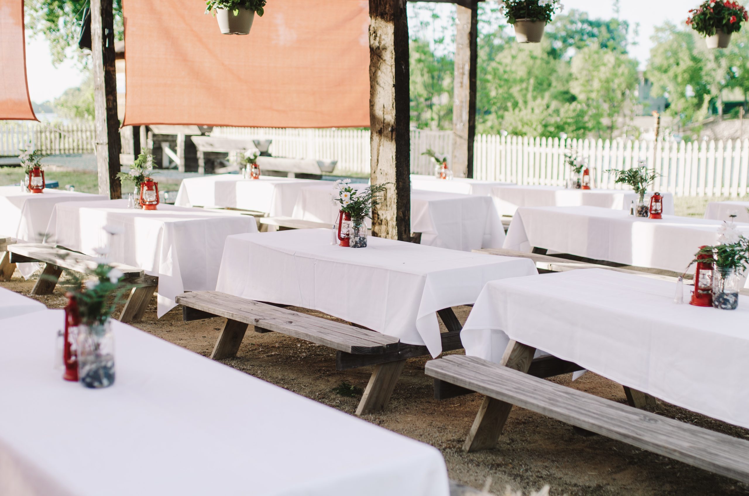 Outdoor picnic tables with white cloths and flower centerpieces under hanging plants.