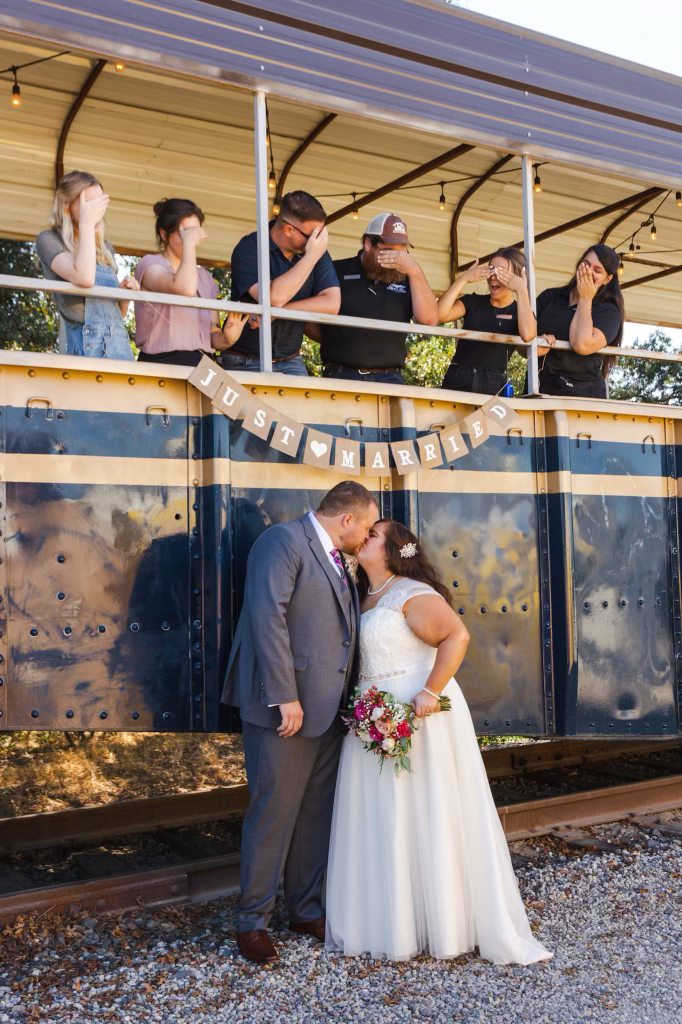 Bride and groom kissing under 'Just Married' banner on train; friends covering eyes above.