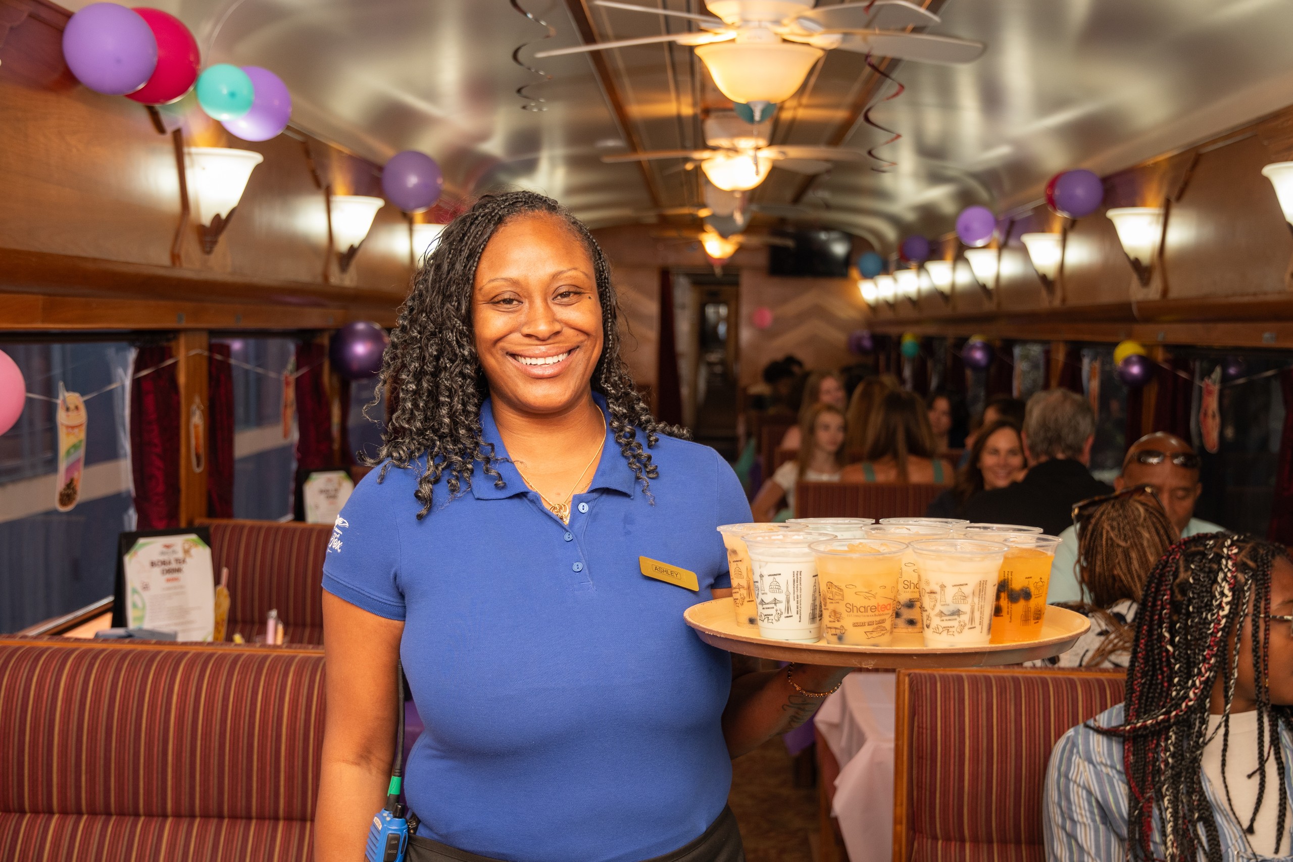 Smiling server holds tray of drinks inside a lively train dining car.