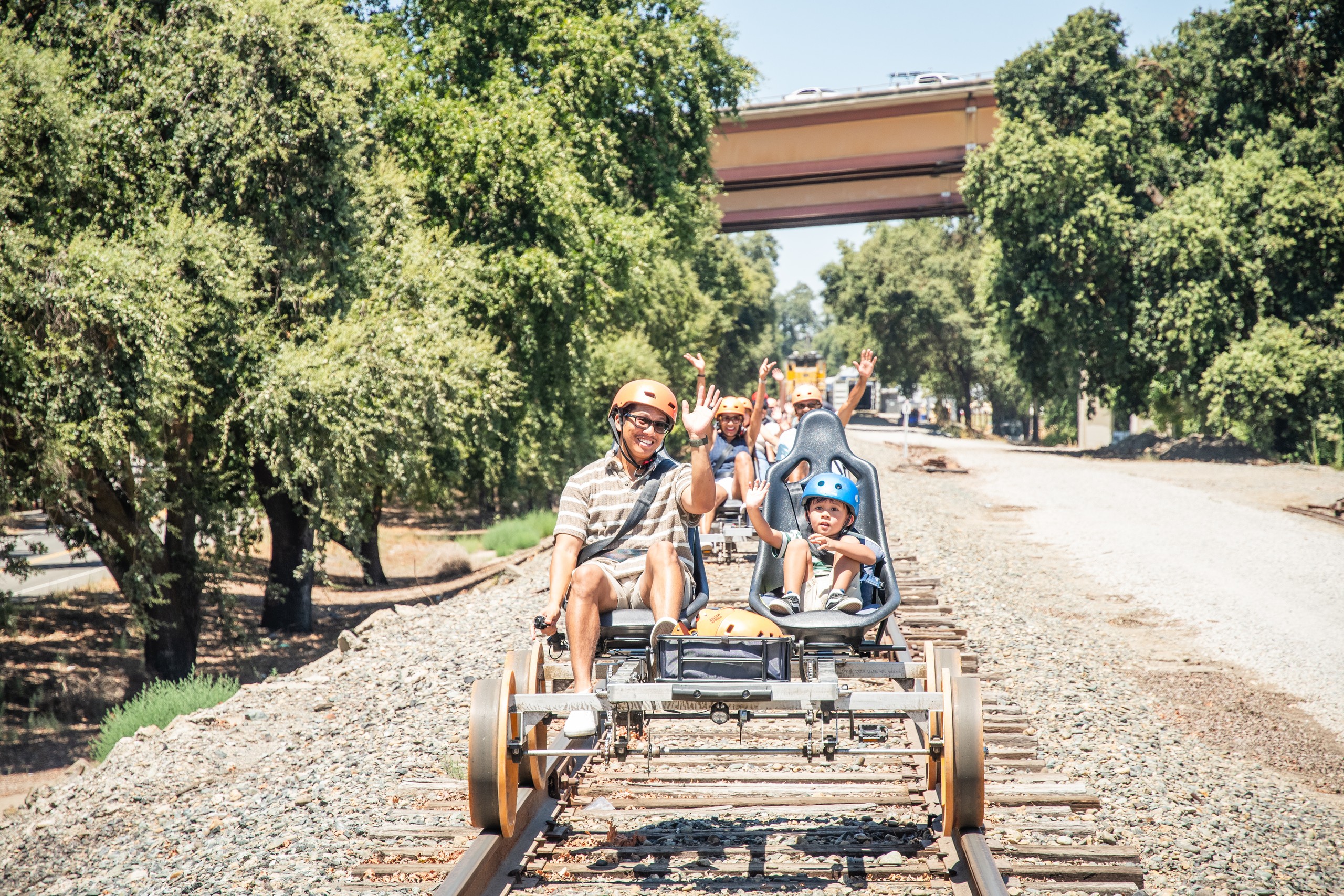 People riding a pedal-powered rail cart on tracks outdoors under trees.