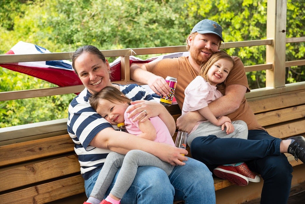 Family of four smiling and sitting on a wooden bench outdoors.